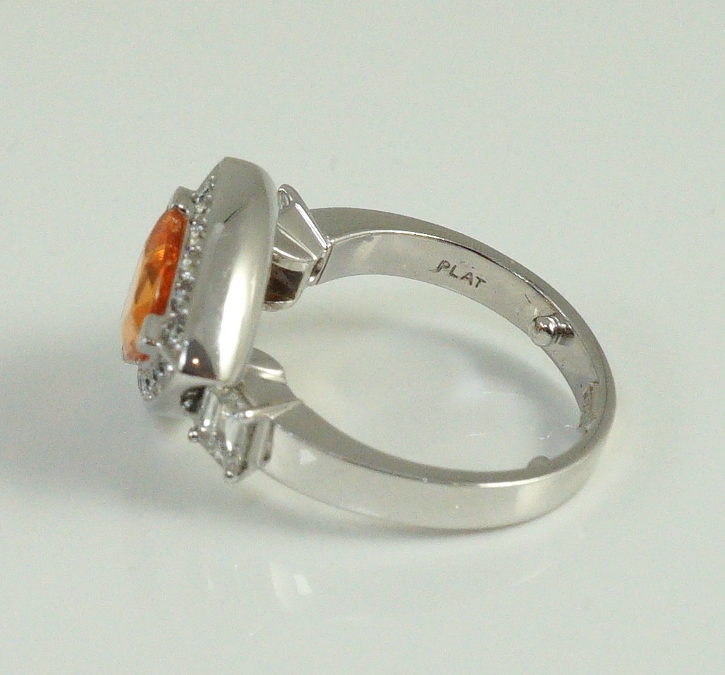 A modern platinum and single stone trilliant cut spessartite garnet ring, with diamond set border and two criss-emerald cut diamond set shoulders, by Musson